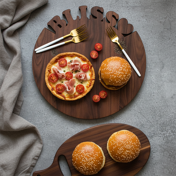 A Personalized Round Cutting Board engraved with "BLISS" holds a small pizza, cherry tomatoes, two forks, and a burger. Next to it, another wooden board displays two sesame seed burgers. A beige cloth sits on the left side.
