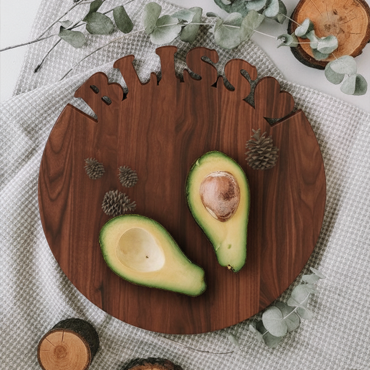 The Personalized Round Cutting Board features "BLISS" cut out at the top, shown with a halved avocado and pinecones, surrounded by eucalyptus leaves and a patterned cloth—ideal for use as a unique charcuterie or wedding board.