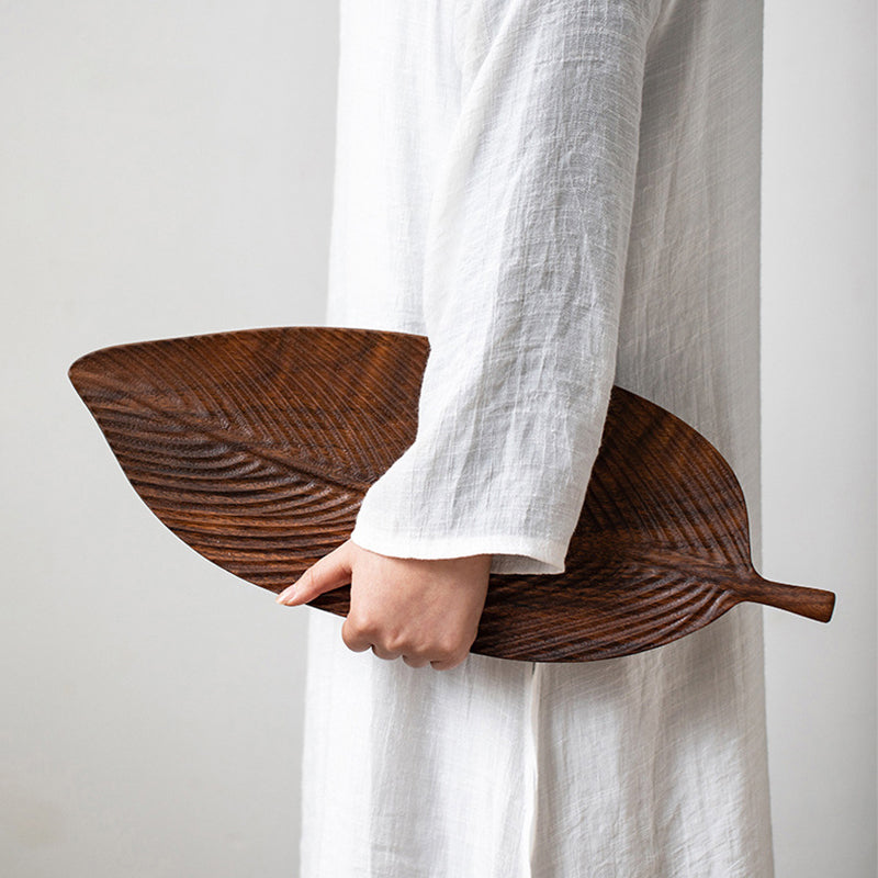 A person in a white long-sleeve top holds a Leaf-Shaped Black Walnut/Rubberwood Serving Tray by their side against a plain background, adding a touch of Japanese-inspired decor.