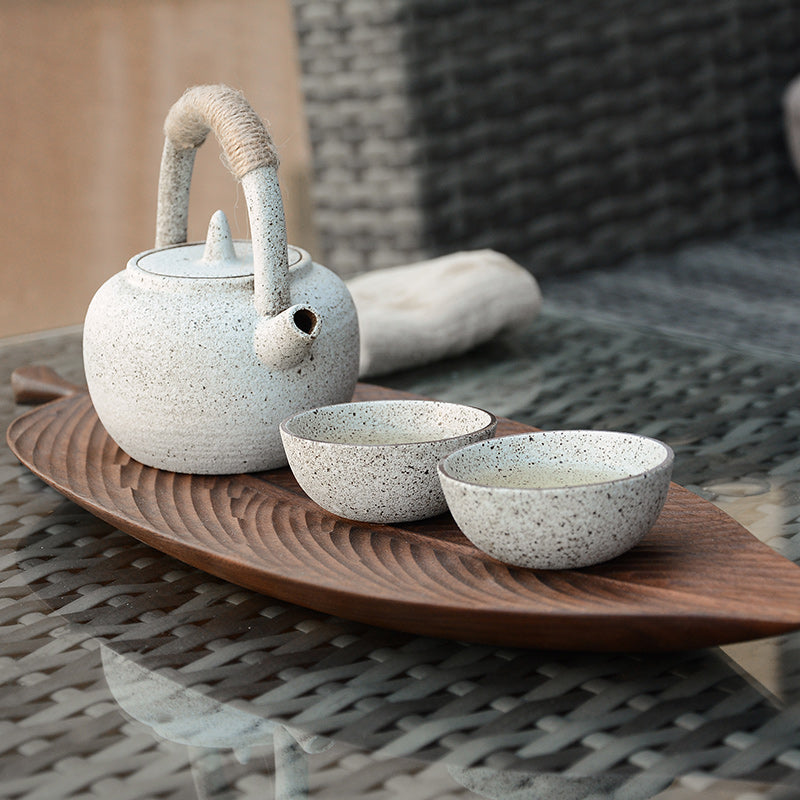 A white speckled ceramic teapot with a wrapped handle and spout sits next to two matching cups on a Leaf-Shaped Black Walnut/Rubberwood Serving Tray atop a glass table with a woven surface underneath.